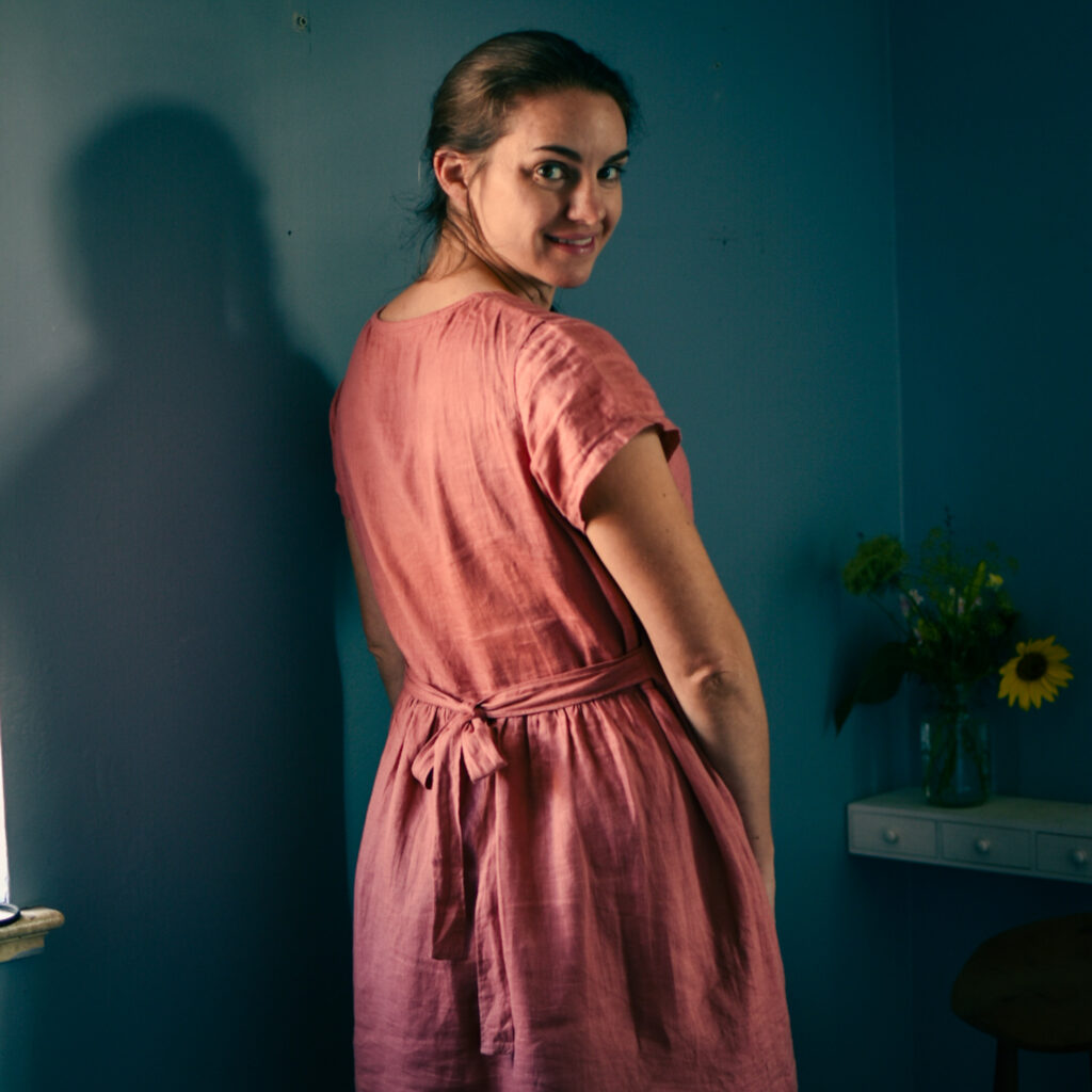 caucasian woman looking over right shoulder at camera in pink, gathered dress with back tie. blue background with vase of flowers