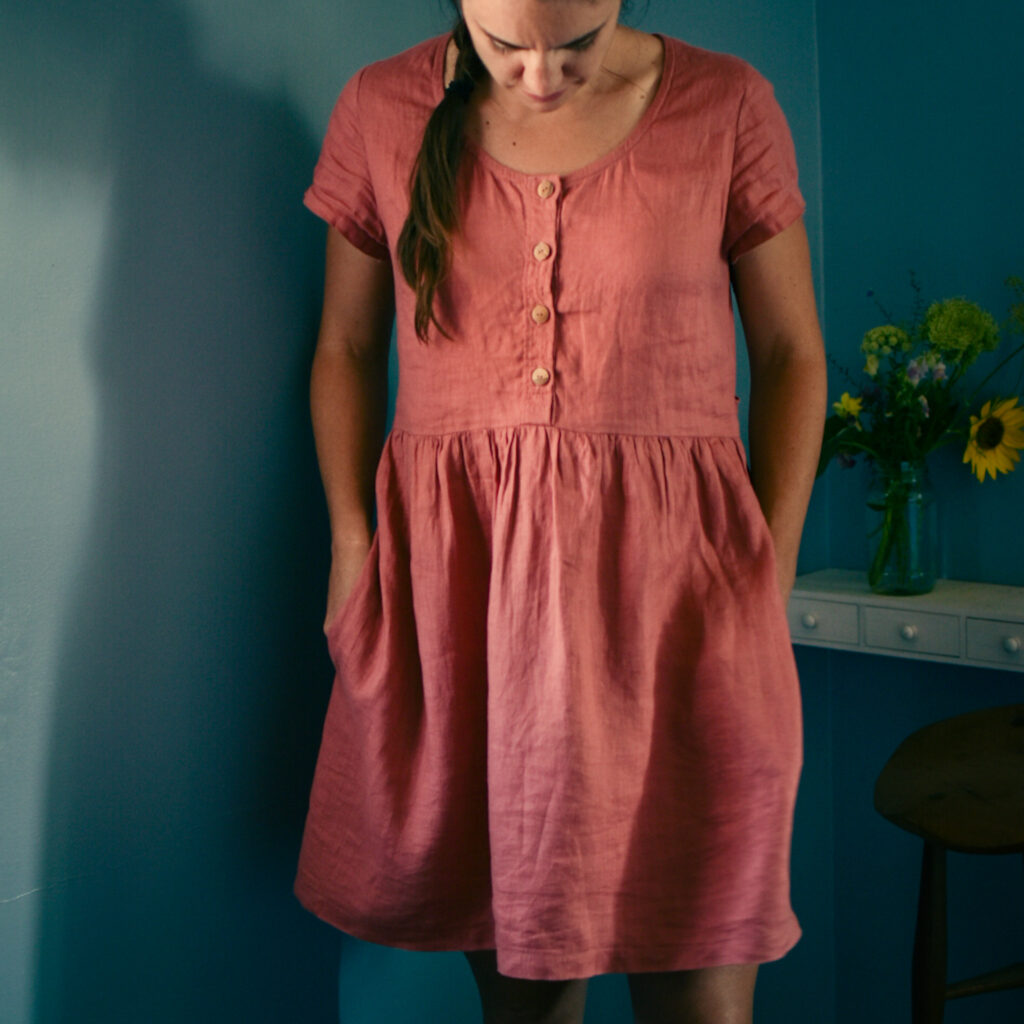 caucasian brown-haired woman staring down at pink, gathered dress with wooden buttons. blue background with flowers