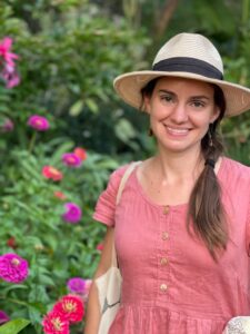 Caucasian female with brown hair, white hat, and pink dress standing in front of pink flowers