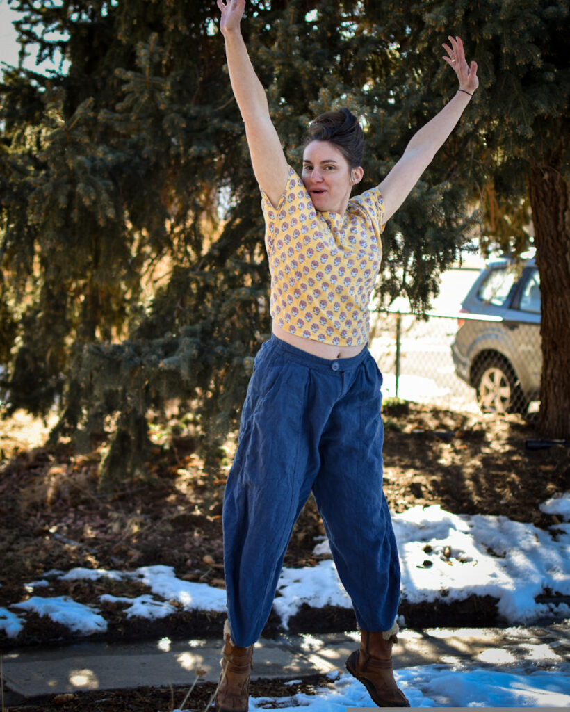 white woman jumping with hands in air. she is wearing high waisted wide legged blue linen pants and floral print yellow cotton baptiste crop top tee shirt