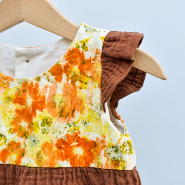 detail of front flowers on 2T toddler dress, sleevless with ruffles, high waist, gathered skirt. bodice is naturally dyed with flowers showing images of sulfur cosmos and marigold petals. skirt and ruffles are brown double gauze cotton with checked texture