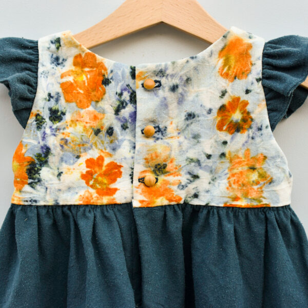 back detail showing 3 wooden buttons of 2T toddler dress, sleevless with ruffles, high waist, gathered skirt. bodice is naturally dyed on raw silk with flowers showing images of sulfur cosmos, scapiosa petals, and purple rose of sharon. skirt and ruffles are an aquamarine raw silk